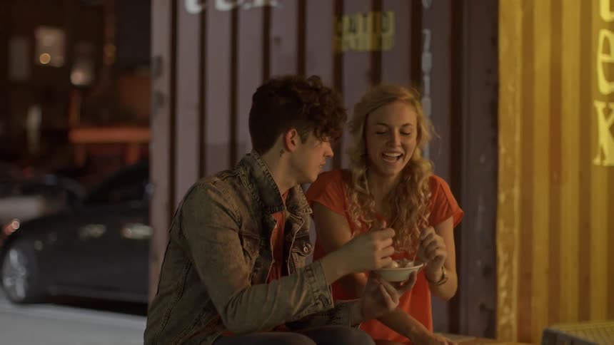 A young playful couple sit on an outside bench sharing a bowl of food