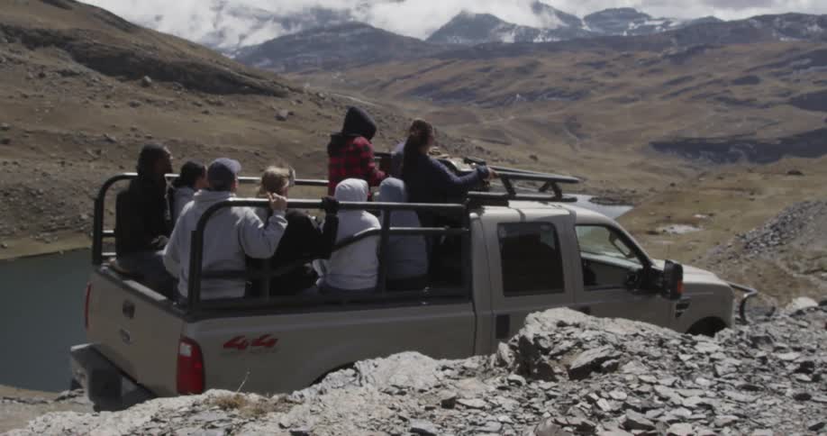 A white pick up truck with people sitting in the bed driving through the rocky terrain near a body of water with a lot of clouds in the sky
