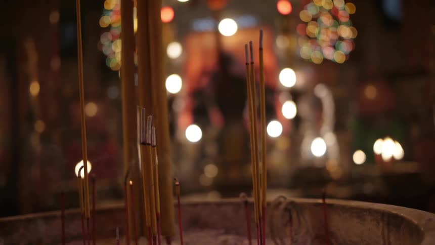 Incense sticks are burning in a large container filled with sand, with candles flickering in the background