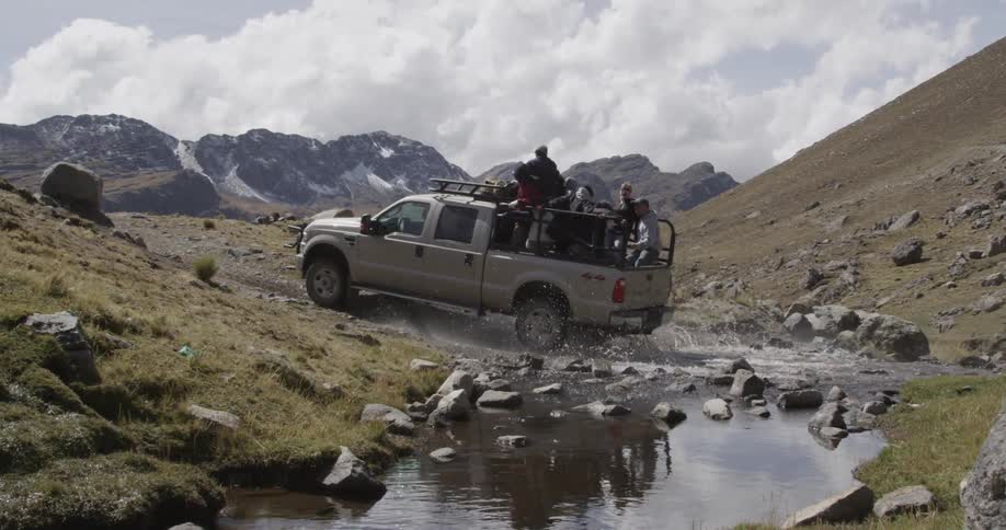 A pickup truck with people in the bed driving through a small stream on rugged terrain