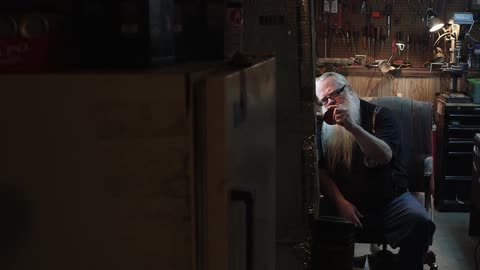 A man with a long white beard sits at a workbench