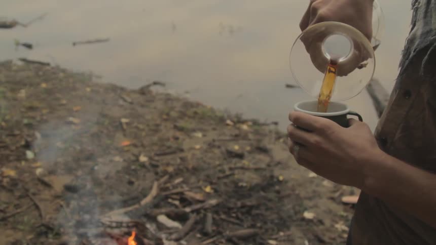A person pours a drink into a mug by a campfire