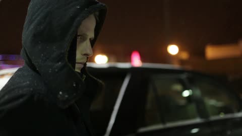 A man with a hooded jacket standing next to a parked car at night