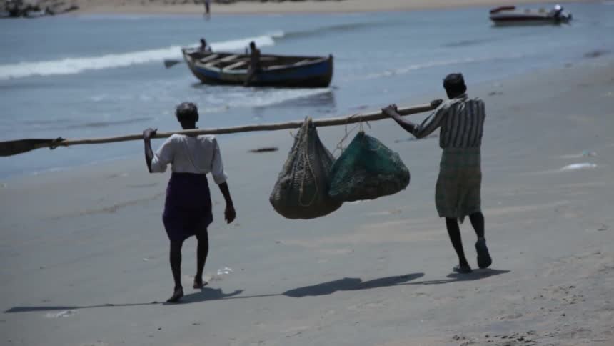 Two people carry bags on a stick on the beach