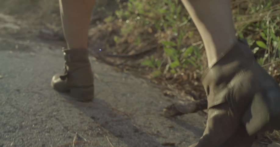 A woman wearing boots walks along muddy ground covered in sticks and tall grass