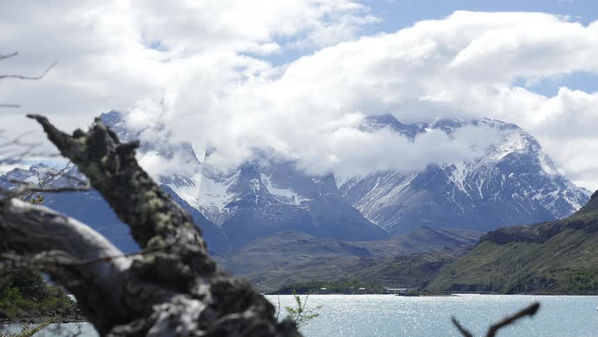 Clouds moving through the bright blue sky above snow covered mountains by a body of water