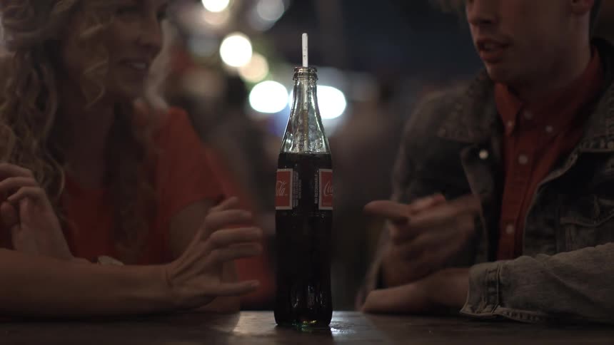 A brunette man and blonde woman seated at a table, talking and laughing with a bottle of coke between them, seemingly having a play argument over who would drink the coke before the woman takes it and sips from the straw