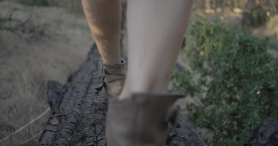 A woman in boots walks across a fallen tree