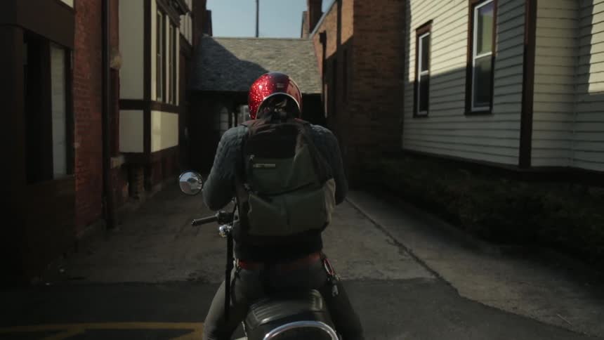A man walks up to his motorcycle, starts it, and rides off down an alleyway
