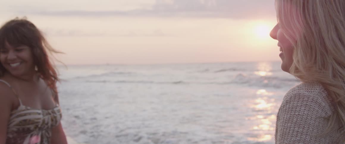 Two women talk and laugh on a beach at dusk