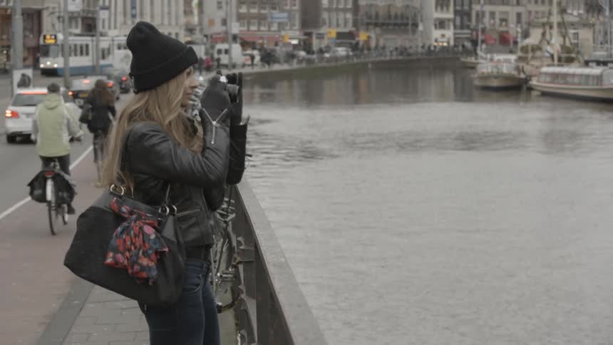 A young, blonde woman standing on a bridge and taking photographs in a city