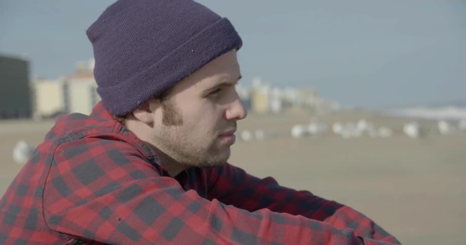 A man in a flannel shirt and hat sitting on a beach during the day