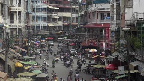 Numerous people riding scooters on a busy city street surrounded by tall buildings and vendor tents