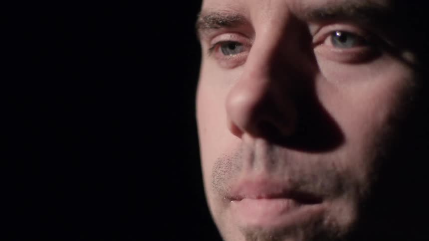 An unshaven, brunette man looking around and appearing to be deep in thought