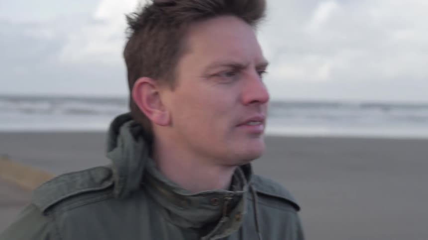 Man standing on a sidewalk alongside a beach, looking around, on a chilly, cloudy day