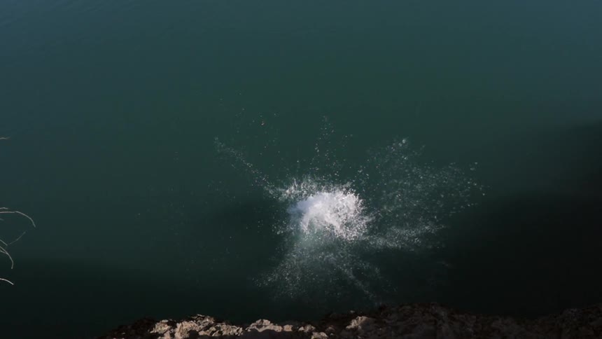 A shirtless man wearing colorful swim trunks dives from a cliff into the water below