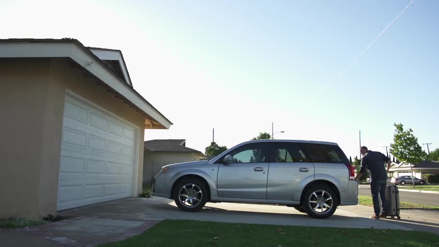 A man rolls a suitcase out to the suv parked in a driveway