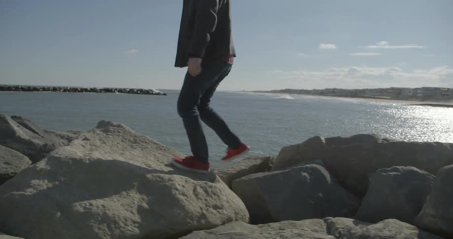A person walking and hopping on top of rocks on the water's edge on a chilly, sunny day