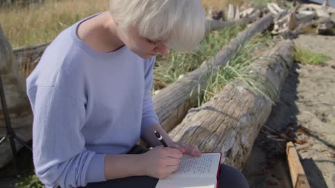 A woman sits on a log and writes in a notebook