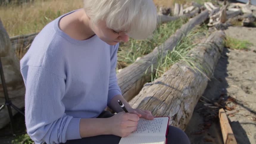 A woman sits on a log and writes in a notebook