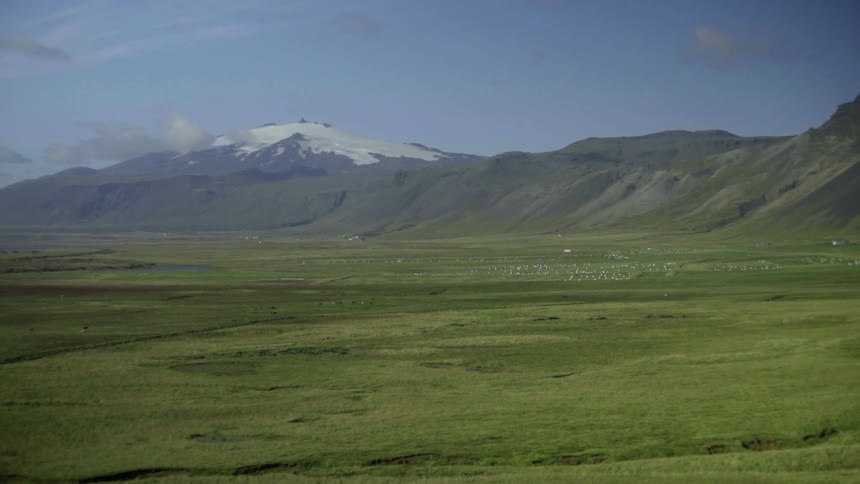 A snow-capped mountain and rolling hills above a flat green valley of grass