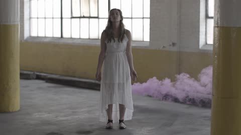 A woman in a white dress standing in a parking garage as purple smoke blows from behind her