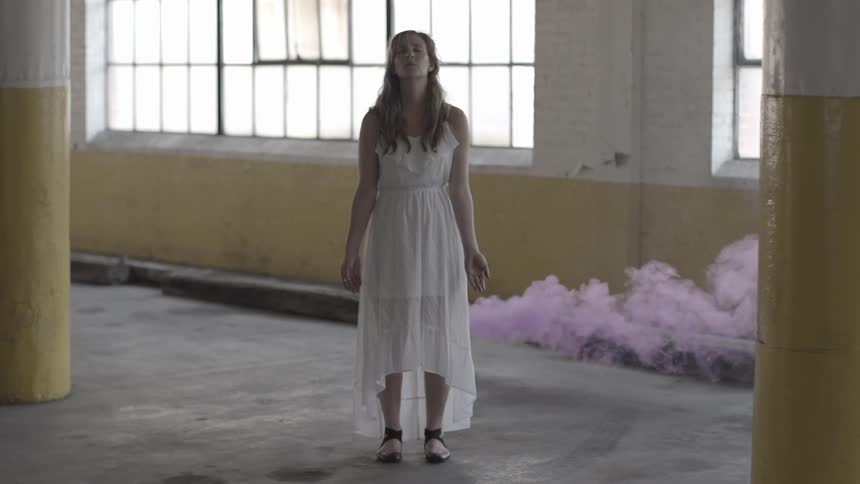 A woman in a white dress standing in a parking garage as purple smoke blows from behind her