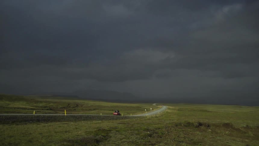 A car driving down a highway heading towards mountains under a sky filled with dark storm clouds