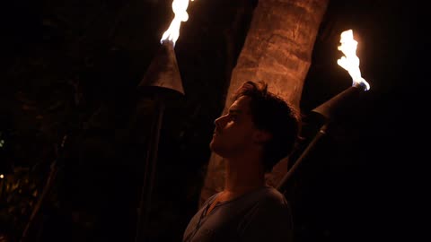 A young, brunette man standing in front of some torches and a palm tree