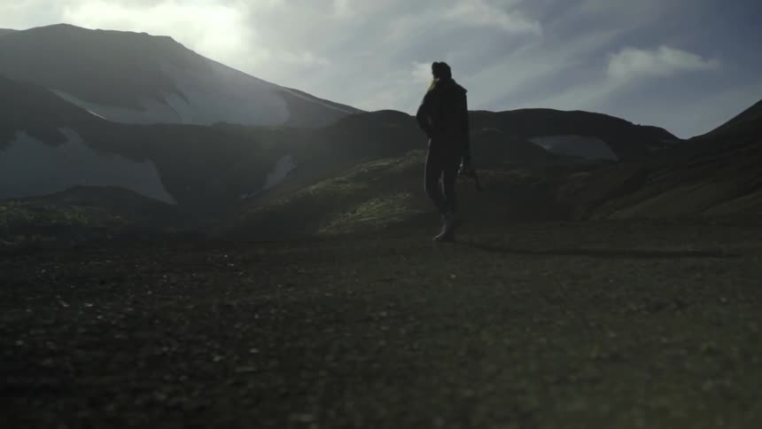 A woman in a knit cap carrying a camera walks in the wilderness looking at a mountain in the distance