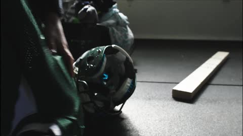 An individual rolling and lifting shoulder pads covered with a football jersey , bumping a football helmet that was resting on the floor