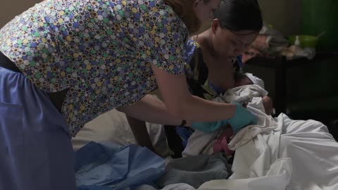 A nurse clamps and cuts the umbilical cord of a newborn as the mother is breast feeding