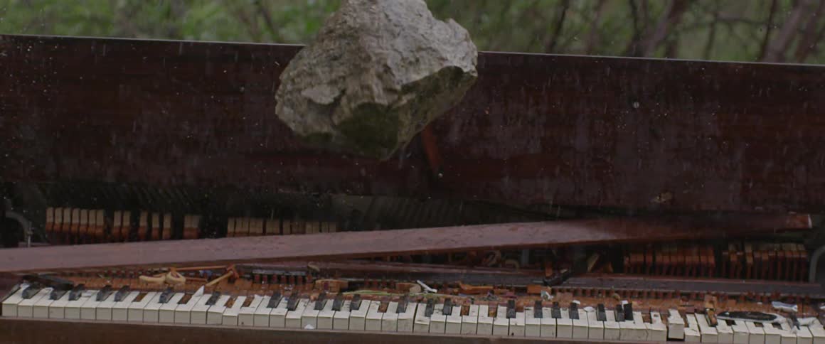 A hand throws a large rock onto a piano outside in the pouring rain