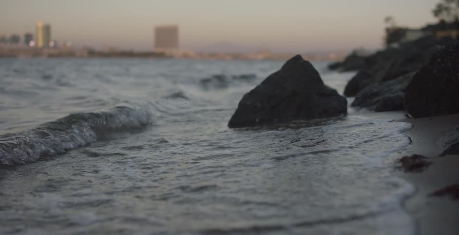 Waves crashing on shore and into rocks in the foreground while several buildings are visible in the background