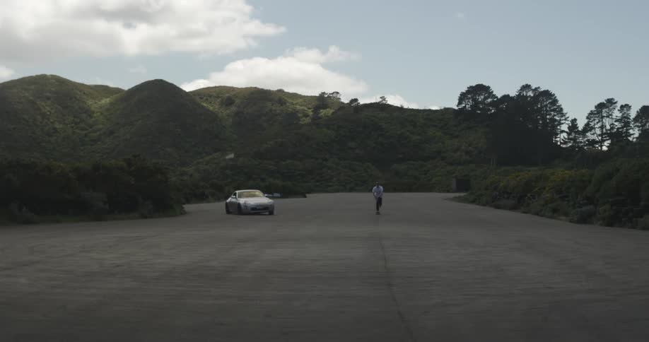 A car races around a man riding a skateboard on a paved lot