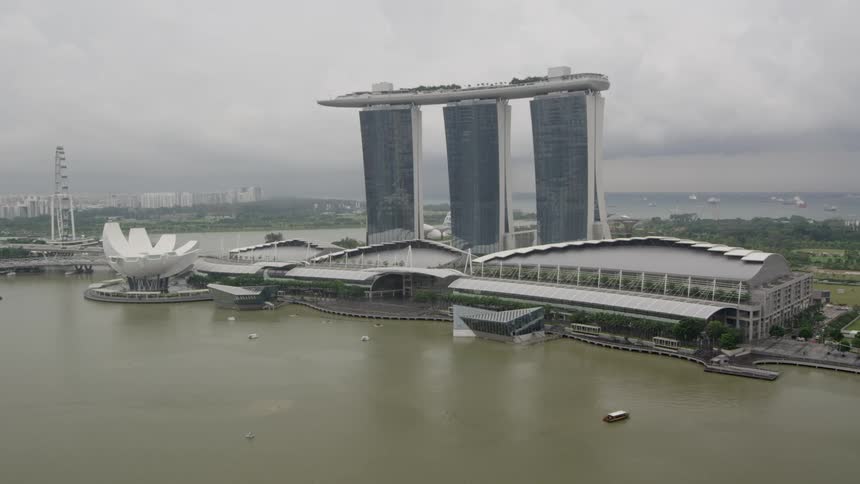A boat moves towards land that has several buildings and three tall towers on it