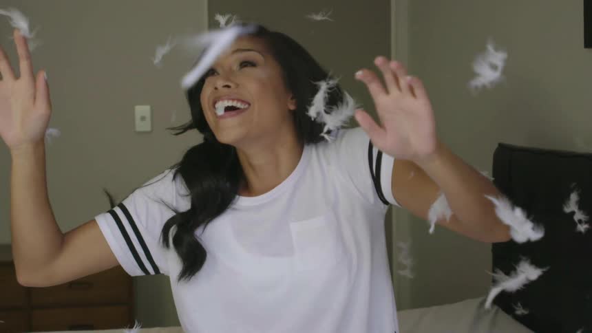 A black-haired woman sitting on a bed, raising her hands and smiling as feathers float around her
