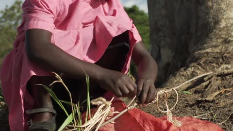 A young girl in a pink dress squatting and wrapping twine around a stick