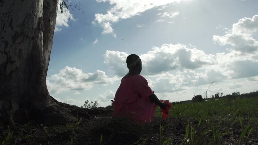 A young girl kneels beside a field near a tree