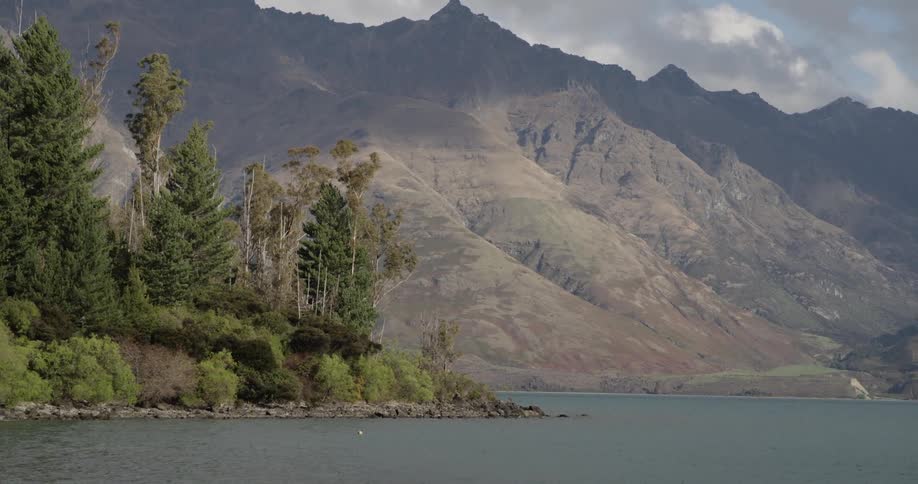 A view of a body of water at the foot of mountains that are partially shaded by dark clouds