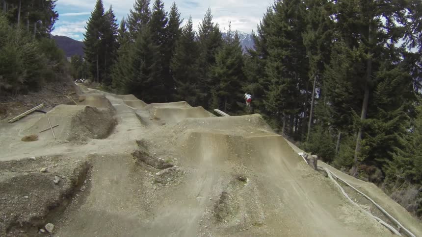 A bicyclist riding a dirt obstacle course surrounded by trees with mountain peaks in the background