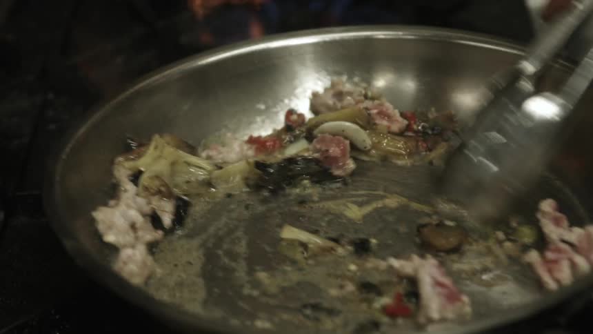 Someone adding spices to a frying pan and stirring the cooking ingredients with a pair of tongs