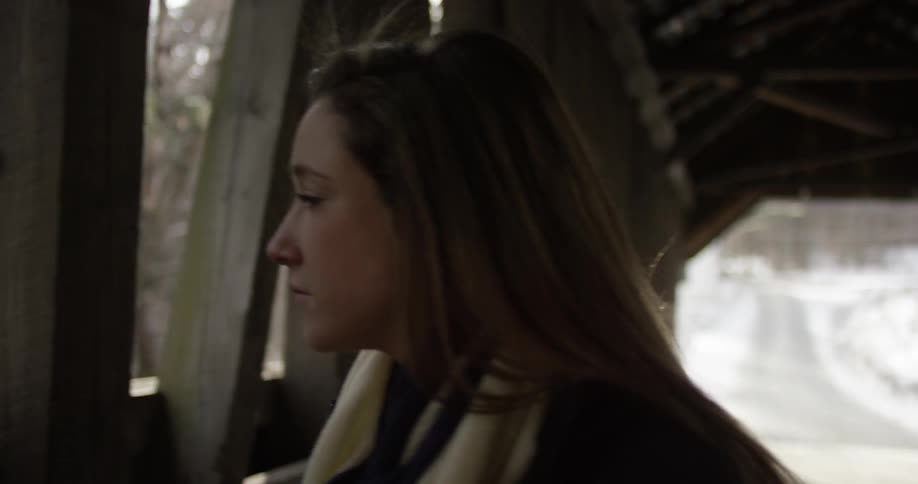 A woman walks across a covered bridge and looks out at something below
