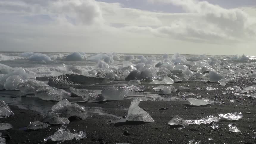 View of chunks of ice that have washed up on a beach as waves crash to shore in the background