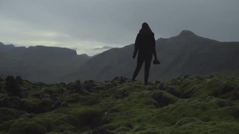 A woman holding a camera in her hand as she stands on top of rocks and surveys the mountainous scene before her