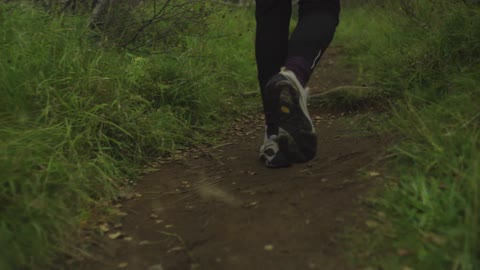A view of a woman's feet as she steps over a large root and walks along a dirt path that is surrounded by tall grass, weeds and brush