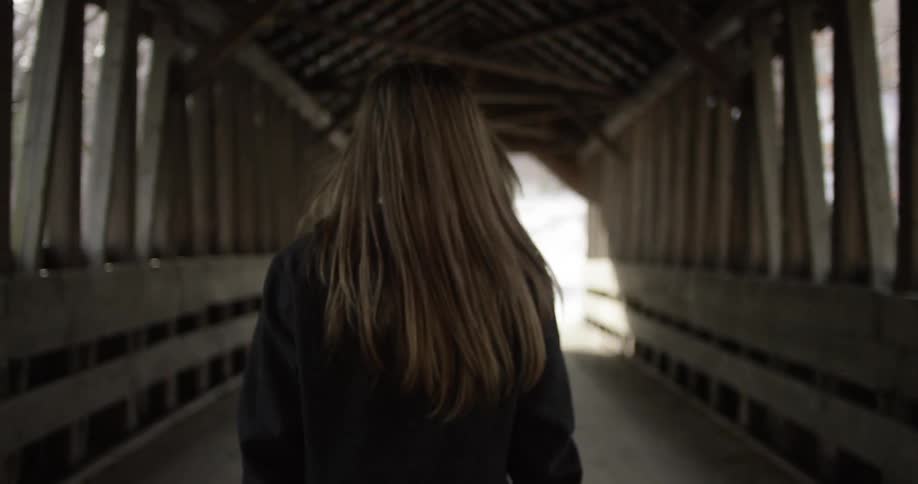 A young woman walks down the center of a covered, wooden bridge