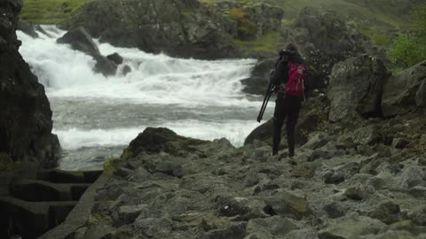 A woman carrying a tripod walking next to white water rapids