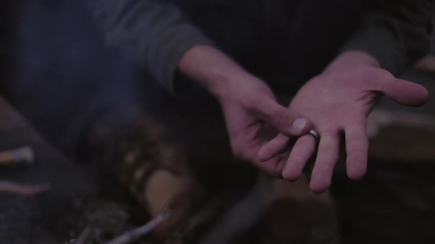 A man sitting by a campfire and twisting his wedding band on his finger