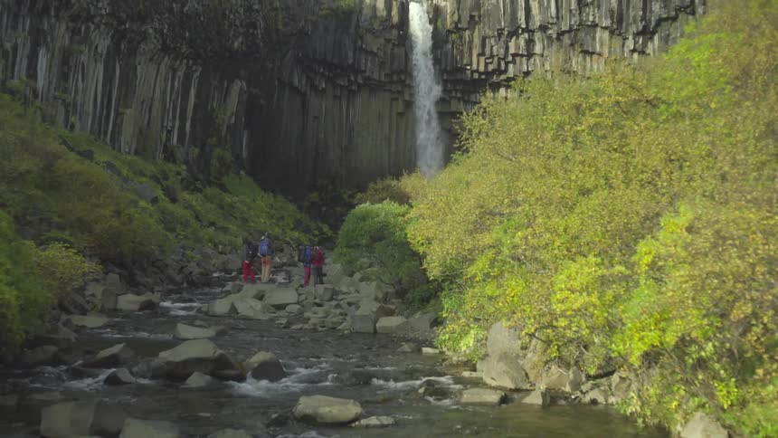 People stand on rocks in a stream near a waterfall in the daytime